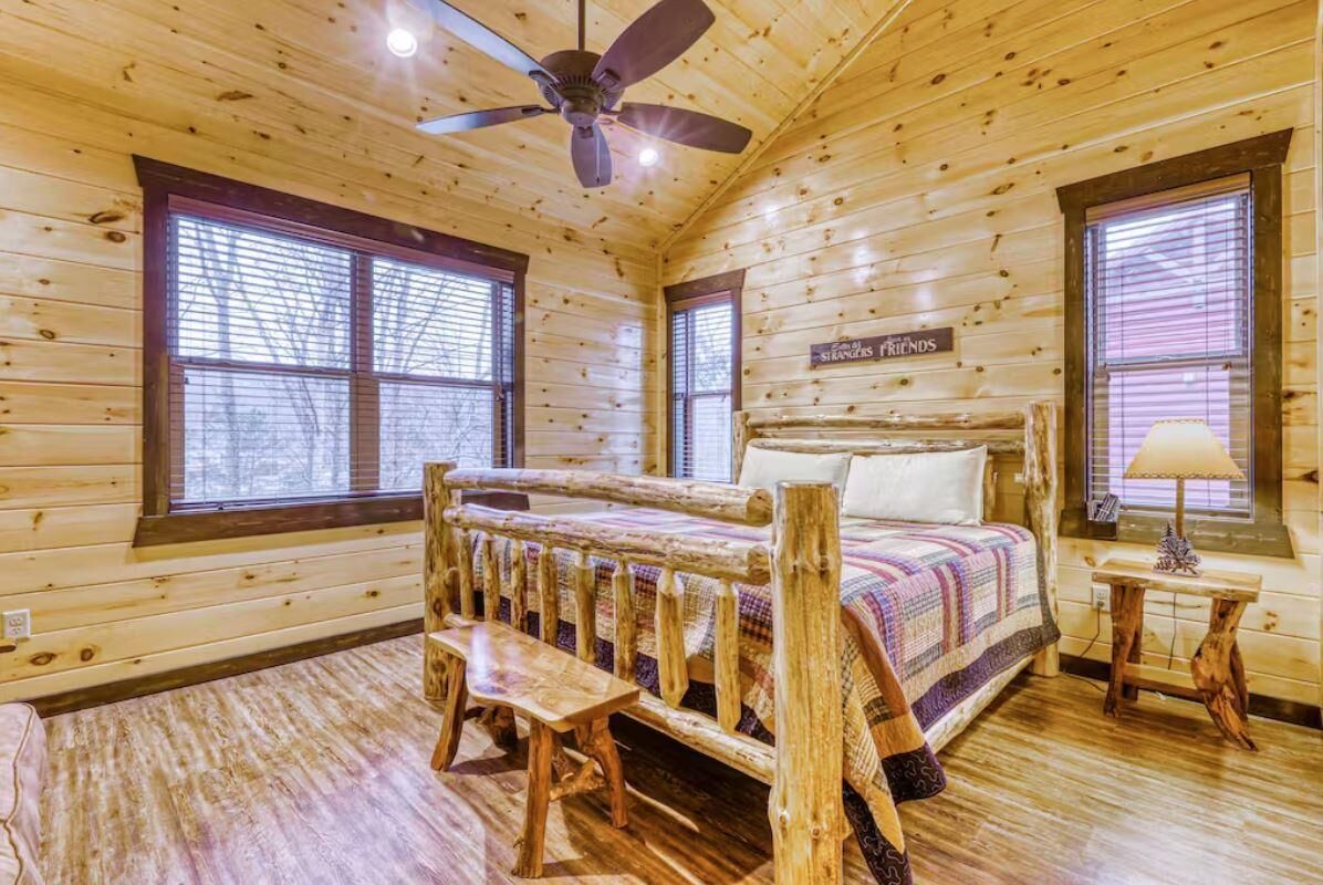 Quarto de cabana de tronco com cama, um banco, ventilador de teto e janela com persianas.