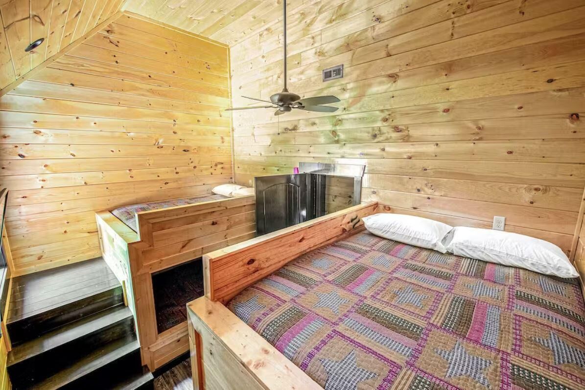 A wooden cabin interior with a bed, a ceiling fan, and a fireplace.