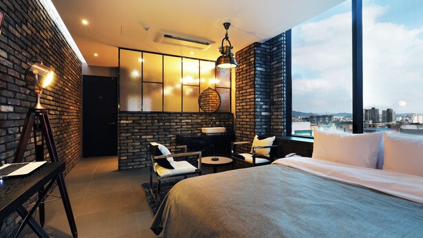 Habitación de hotel moderna con una cama grande, un escritorio con una computadora portátil, una zona de estar con sillas, una mesita y vistas a la ciudad a través de amplios ventanales.
