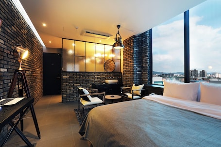 A modern hotel room with a large bed, a desk with a laptop, a seating area with chairs, a small table, and a city view through large windows.. Cheonan LaTree Hotel