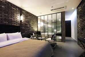 A modern hotel room with a brick wall, a bed, a seating area with a table, and a sliding glass door leading to a balcony.