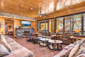 A cozy lodge interior with a stone fireplace, wooden ceiling, and large windows offering a view of autumn foliage. - The Grand Orchard Lodge (Gatlinburg)