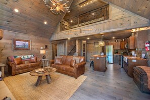 A spacious living area with a high ceiling, wooden walls, a fireplace, and a kitchen in the background. - The Aspen Cabin (Gatlinburg)