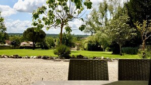 A gravel patio with wicker furniture, a tree, and a view of a green landscape with houses in the background. - 3-star air-con gîte - 5 mins by foot to Monpazier at the heart of the bastides. (Capdrot)