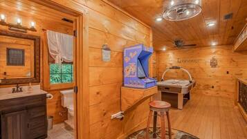 Interior de una cabaña de madera con tocador, lavabo, espejo, ventana con cortinas, ventilador de techo y una máquina arcade azul.