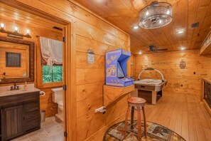 Interior de uma cabana de madeira com toucador de madeira, pia, espelho, janela com cortinas, ventoinha no teto e máquina de jogos eletrônicos azul.