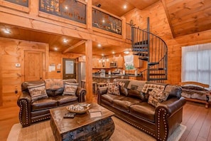 A wooden cabin interior with two leather sofas, a coffee table, a spiral staircase, and a kitchen area. - The Buckhorn (Gatlinburg)