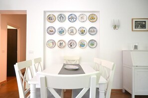 Een eetgedeelte met een witte tafel en stoelen, een wandcollectie borden en een klein kastje.