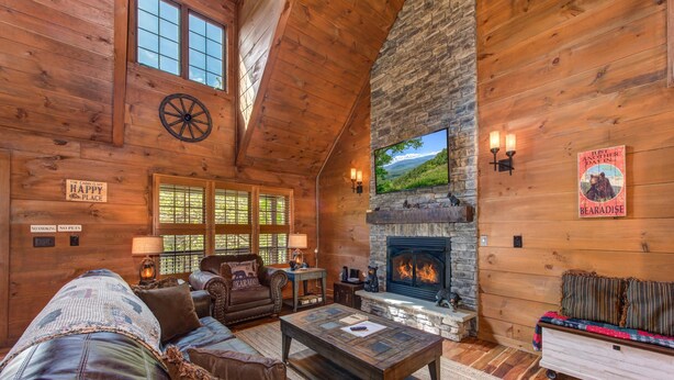 A cozy cabin interior with a stone fireplace, wooden walls, a leather sofa, and a television mounted above the fireplace. - Black Bear Cabin (Gatlinburg)