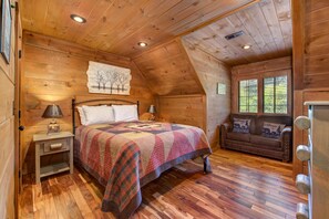 Un dormitorio de cabaña de madera con cama, mesita de noche, lámpara y un sofá con vistas al exterior.