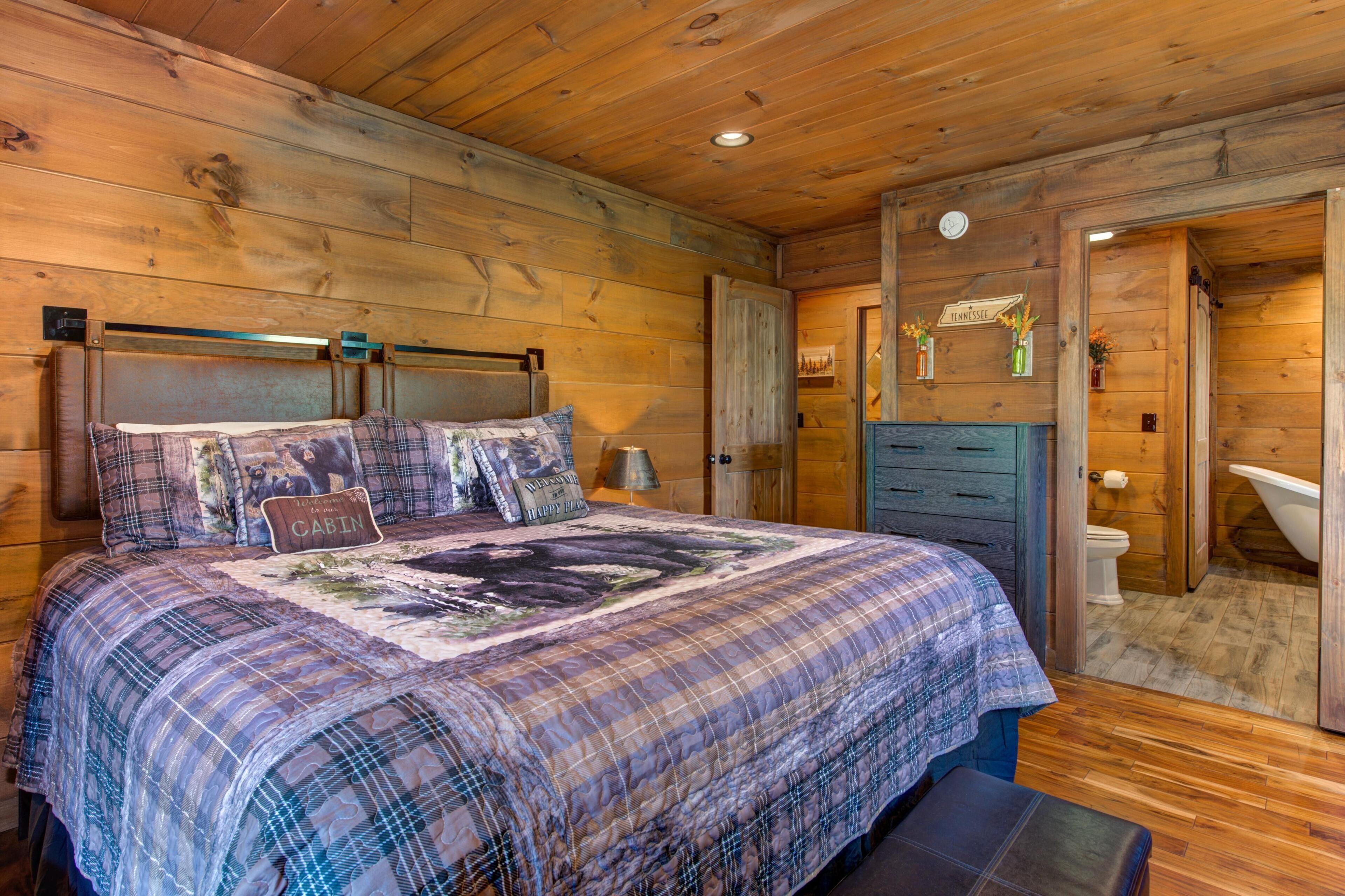 Una habitación tipo cabaña con una cama, techo y pared de madera, baño con inodoro y lavabo, y una puerta que da a otra habitación.