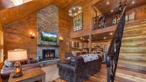 A log cabin interior with a fireplace, wooden floors, a sofa, a dining table, and a staircase. - Black Bear Cabin (Gatlinburg)