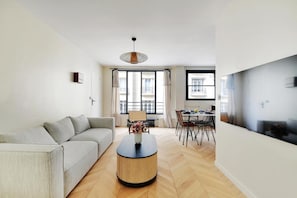 Ein modernes Wohnzimmer mit einer Couch, einem runden Couchtisch, einem Essbereich mit Stühlen und einem großen Fenster.