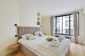 Ein Hotelzimmer mit einem Bett, einem kleinen Tisch, einem Fenster mit Vorhängen und Blick auf Gebäude.