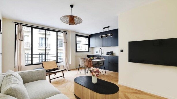 Una sala moderna con un sofá, una mesa de comedor con sillas, una mesa de centro con un florero, y una zona de cocina con armarios y un televisor.