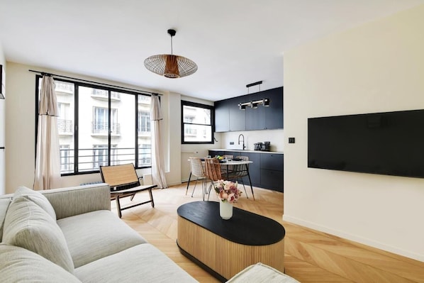 Ein modernes Wohnzimmer mit Sofa, Esstisch und Stühlen, Couchtisch mit Vase sowie Küchenzeile mit Schränken und Fernseher.