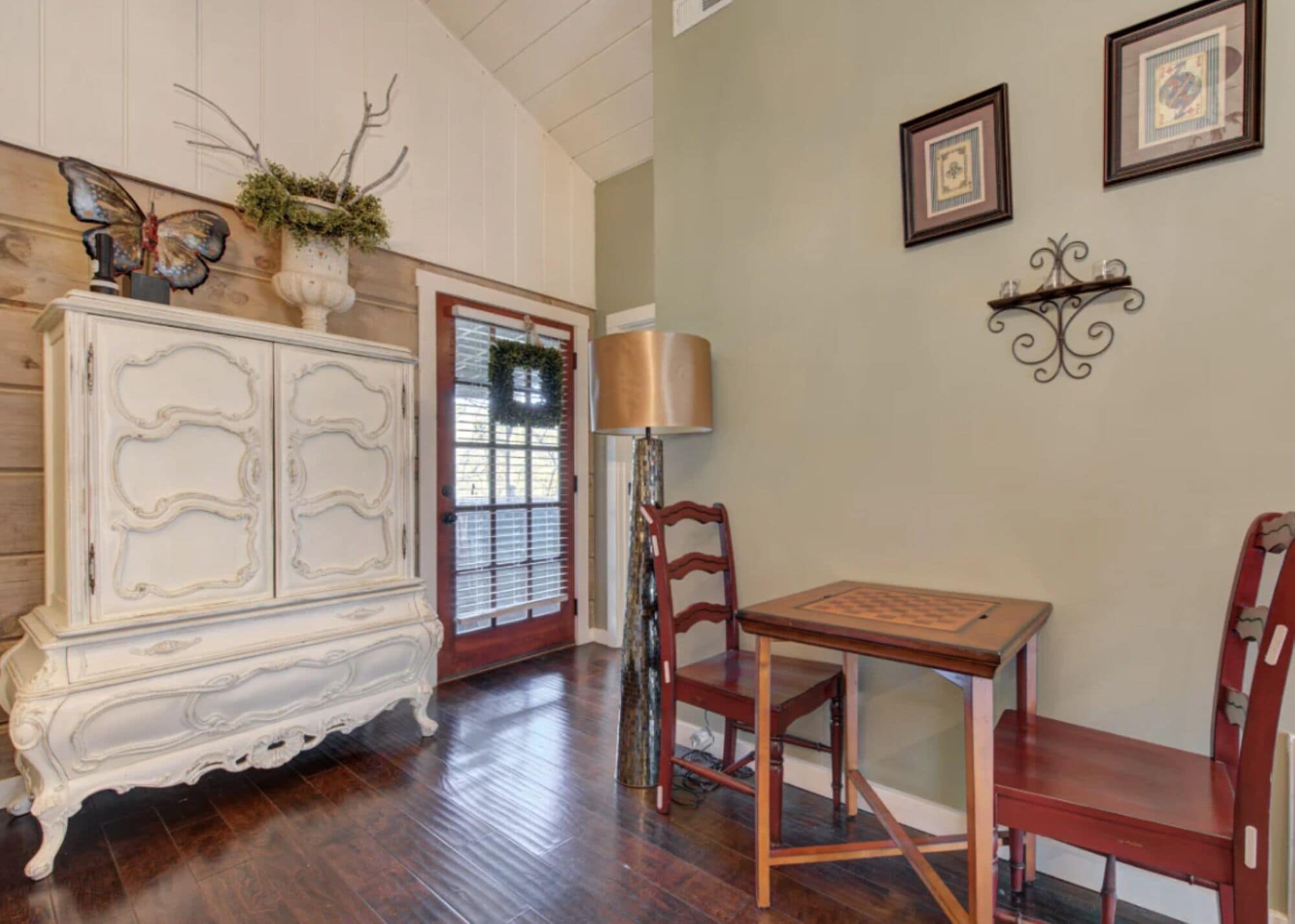 A room with a wooden floor, a white ornate cabinet, a wooden table with chairs, a door with a wreath, and wall-mounted pictures.