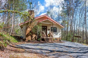 Une cabane avec une cheminée en pierre, une véranda en bois et une allée de gravier, le tout entouré d’arbres.