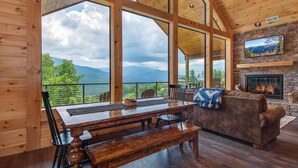 Un interior de madera con una mesa de comedor, sillas, un sofá, una chimenea y un gran ventanal con vistas a un paisaje montañoso.