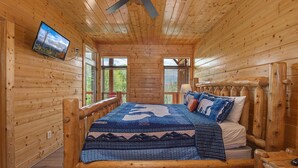 A wooden cabin room with a bed, a ceiling fan, a TV mounted on the wall, and windows. - Papa Bears Magnificent View (Gatlinburg)