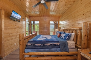 Ein Zimmer in einer Holzhütte mit einem Bett, einem Deckenventilator, einem an der Wand befestigten Fernseher und Fenstern.