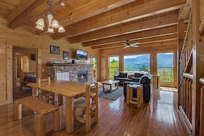 Innenansicht einer Holzhütte mit Kamin aus Stein, Holzmöbeln, einem Fernseher und großen Fenstern mit Blick auf die Berge.