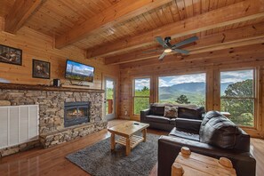 Ein Holzinnenraum mit Kamin aus Stein, einem Fernseher, einer Couch und großen Fenstern mit Blick auf die Berge.