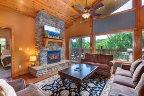A cozy living room with a stone fireplace, comfortable seating, and large windows overlooking a deck. - Golden Oak Lodge (Gatlinburg)