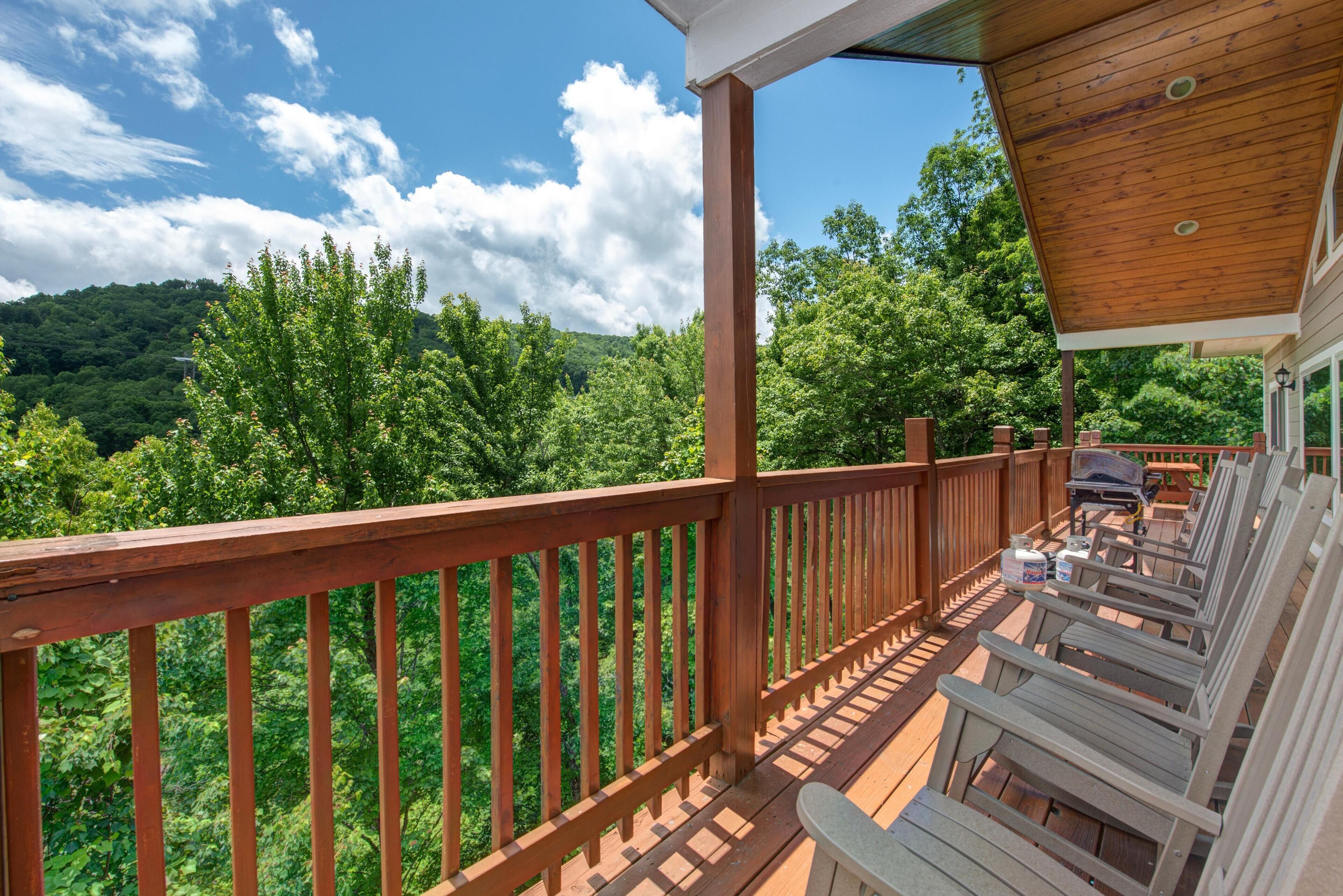 A wooden deck with a view of a forested hillside, outdoor seating, and a barbecue grill.