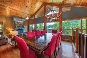 A spacious dining area with a large wooden table and red upholstered chairs, a stone fireplace, and a view of the outdoors through large windows. - Golden Oak Lodge (Gatlinburg)