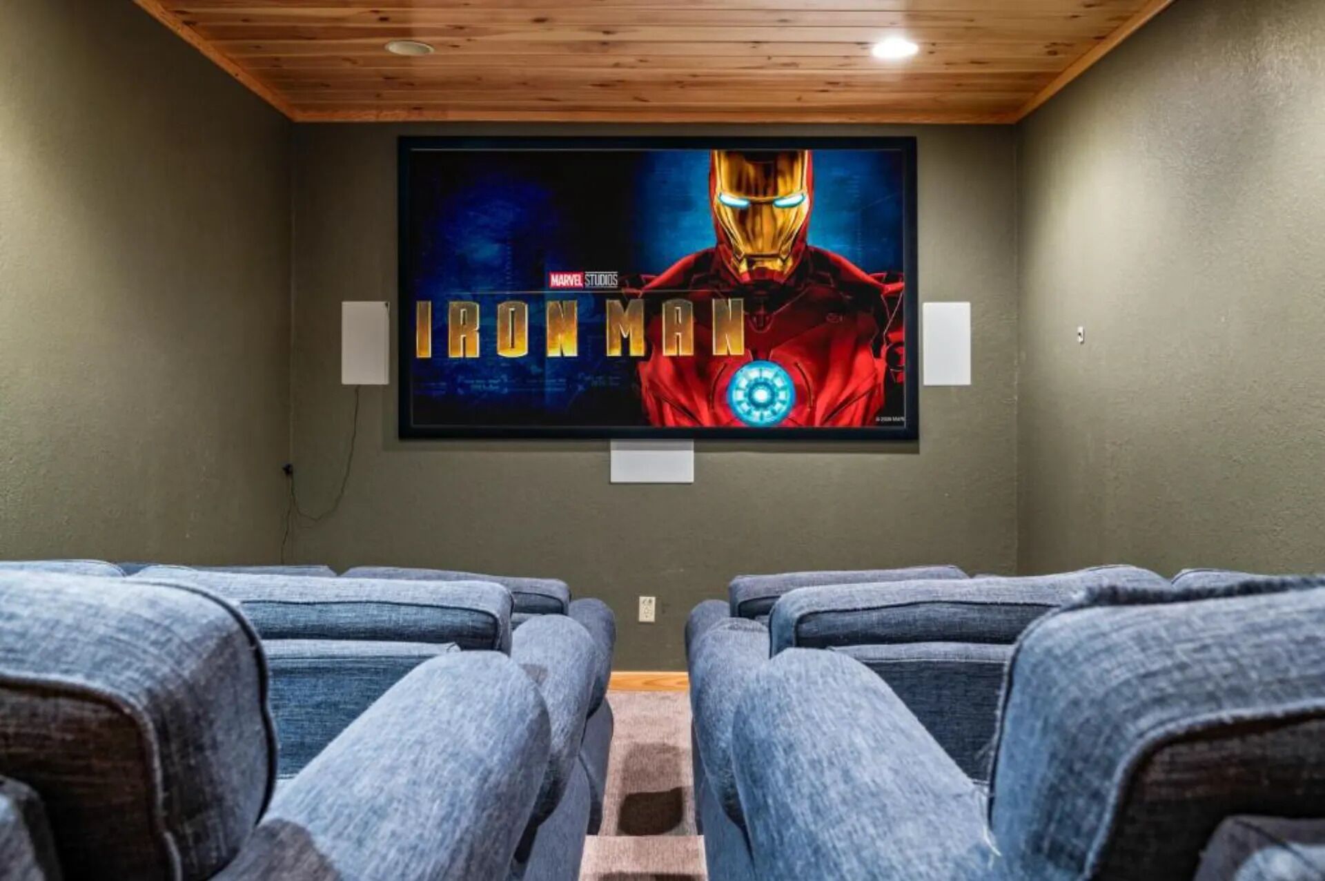 A home theater with a large screen displaying an Iron Man movie poster, two rows of seating, and a wooden ceiling.