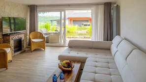 Ein Wohnzimmer mit Kamin, einer Couch, einem Fernseher und Blick auf einen Garten sowie ein Gebäude.