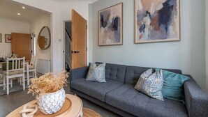 Ein modernes Wohnzimmer mit grauem Sofa, dekorativen Kissen, abstrakter Wandkunst, einem Couchtisch mit einer Vase und einem Essbereich im Hintergrund.
