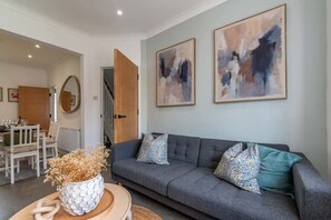 Ein modernes Wohnzimmer mit grauem Sofa, dekorativen Kissen, abstrakter Wandkunst, einem Couchtisch mit einer Vase und einem Essbereich im Hintergrund.