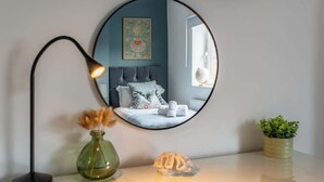Ein runder Spiegel spiegelt ein Schlafzimmer mit Bett, Kissen, einer Weltkugel und einer Vase mit getrockneten Blumen.