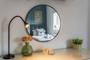 Ein runder Spiegel spiegelt ein Schlafzimmer mit Bett, Kissen, einer Weltkugel und einer Vase mit getrockneten Blumen.