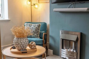 Ein gemütliches Wohnzimmer mit einem blauen Sofa, einem gemusterten Zierkissen, einer Dekovase, einem Kamin und einem an der Wand montierten Fernseher.