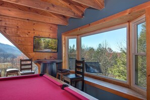 A pool table in a room with large windows, wooden ceiling beams, and a view of trees and mountains. - Honey Bear (Gatlinburg)