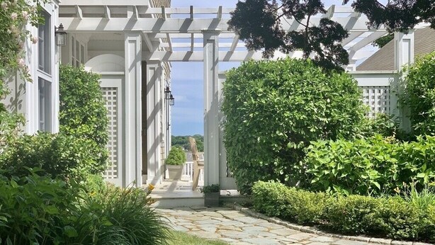 En gång leder fram till en vit pergola, och från den har man utsikt över en avlägsen byggnad.