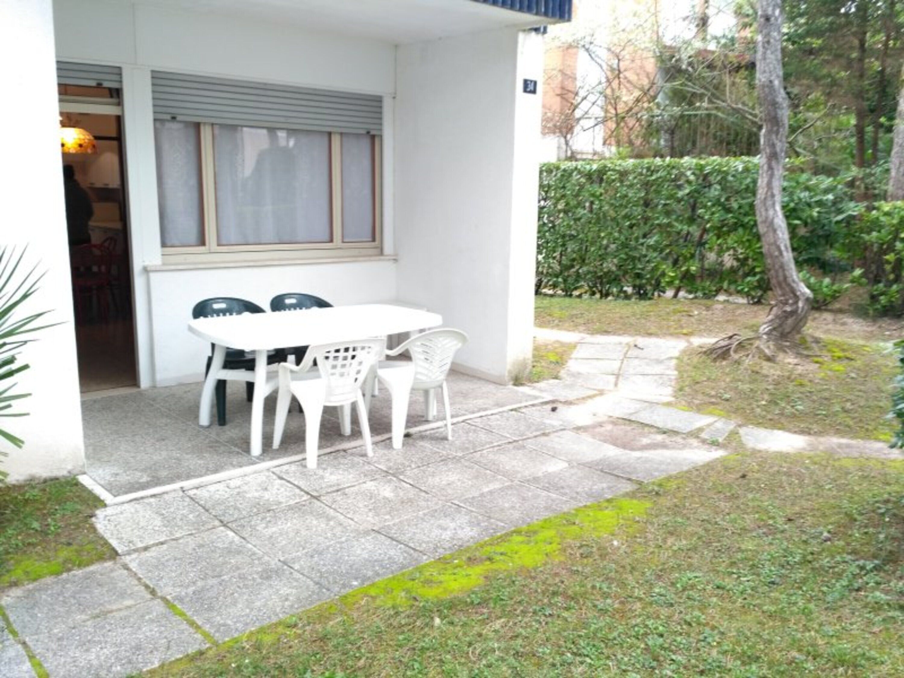 A patio with a white table and chairs, a window with blinds, and a hedge in the background.