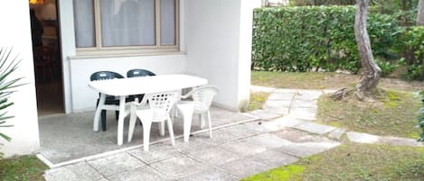 Un patio con una mesa y sillas blancas, una ventana con persianas, y un seto al fondo.