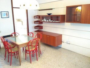 Ein Essbereich mit einem Glastisch und roten Stühlen, einem Holzschrank mit Regalen sowie einem an der Wand befestigten Regal mit Dekoration.