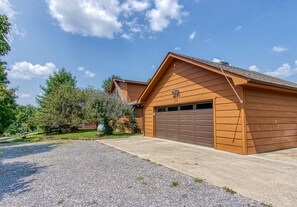 Eine zweistöckige Holzhütte mit Garage, umgeben von einem Schotterhof und viel Grün.