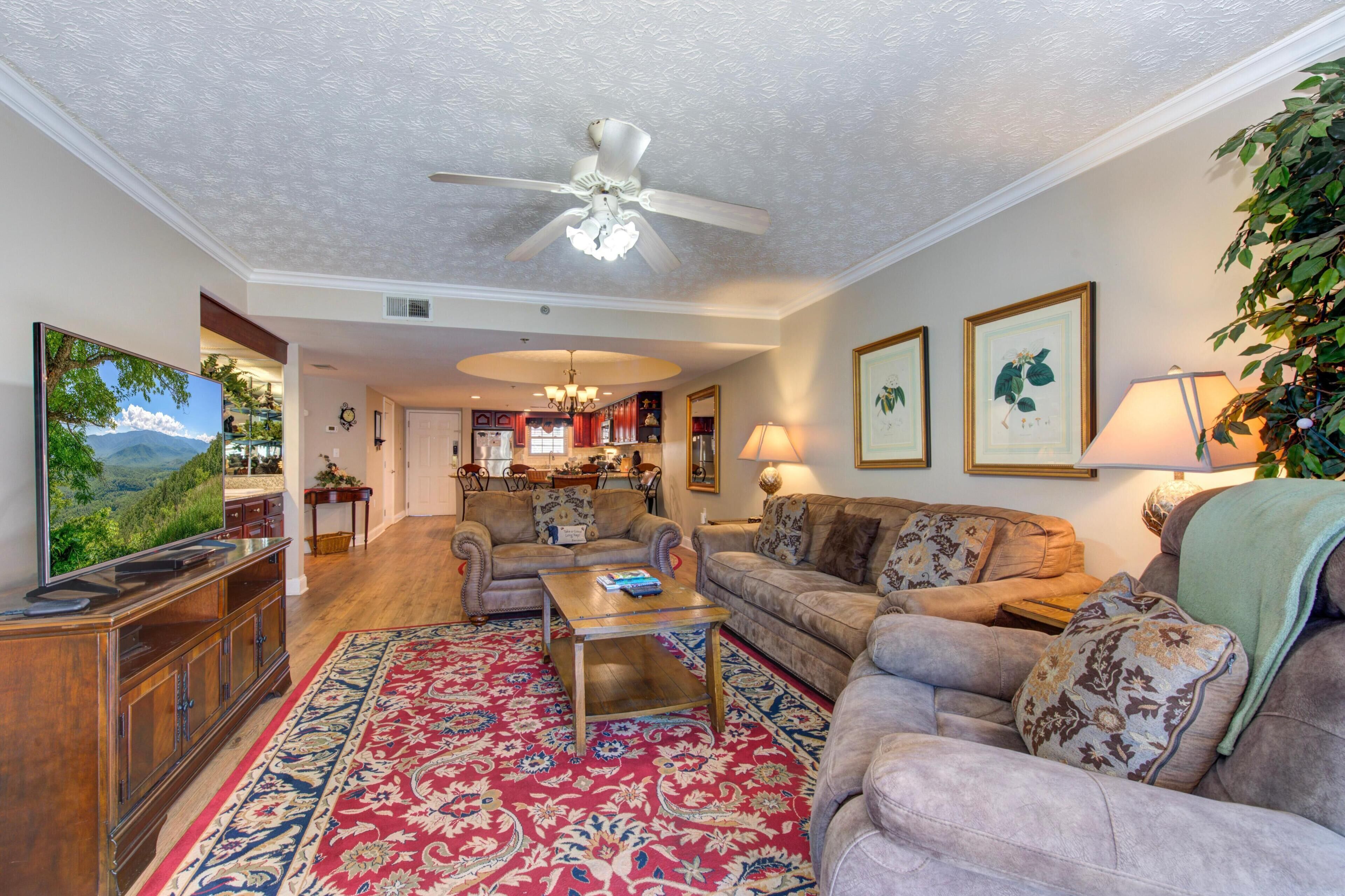 A spacious living room with a ceiling fan, a large flat-screen TV, a wooden coffee table, and a red patterned rug.