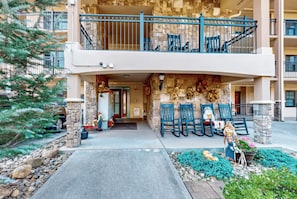Un edificio con fachada de piedra, un balcón con barandas azules y mobiliario exterior con sillas y una figura decorativa.