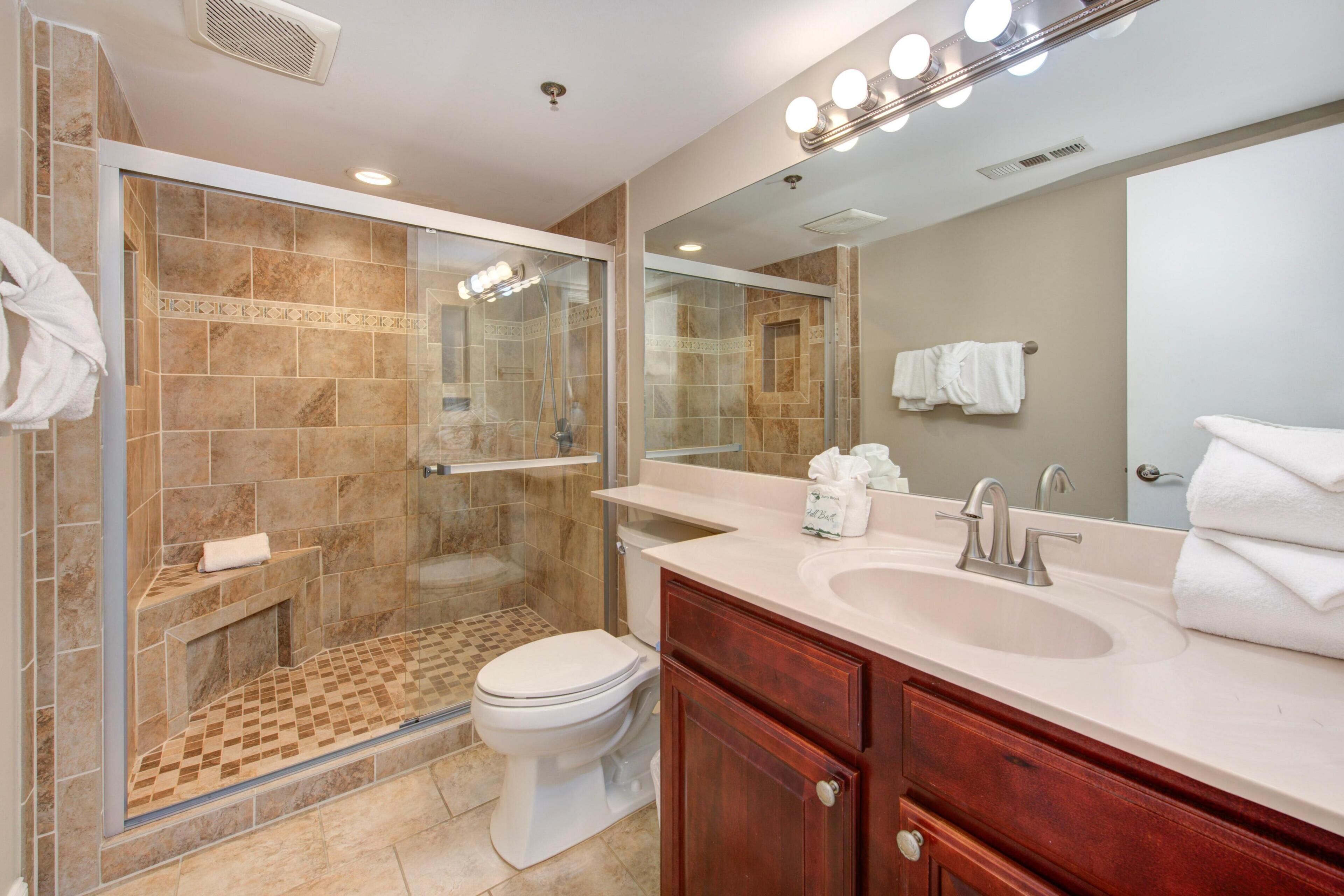 Un baño moderno con ducha de vidrio, inodoro, lavabo con tocador y un espejo grande con luces.