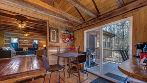 A wooden cabin interior with a dining area, a sofa set, and a large sliding glass door leading to an outdoor deck. - Cricket Hollow Lodge (Cosby)