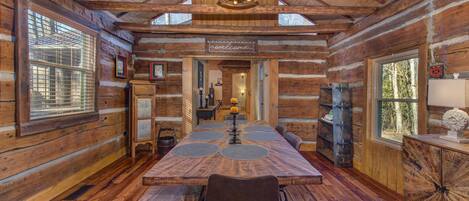 A log cabin interior with a wooden dining table, chairs, a chandelier, and a 'welcome' sign.