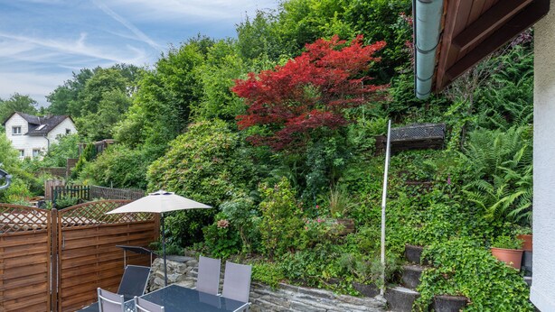 Une terrasse avec une table et des chaises, entourée d’une végétation luxuriante et d’un arbre aux feuilles rouges.