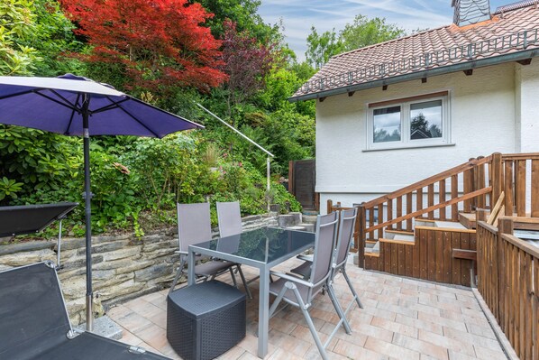 A patio with a glass table and chairs, a purple umbrella, and a wooden staircase leading to a white building with a tiled roof.